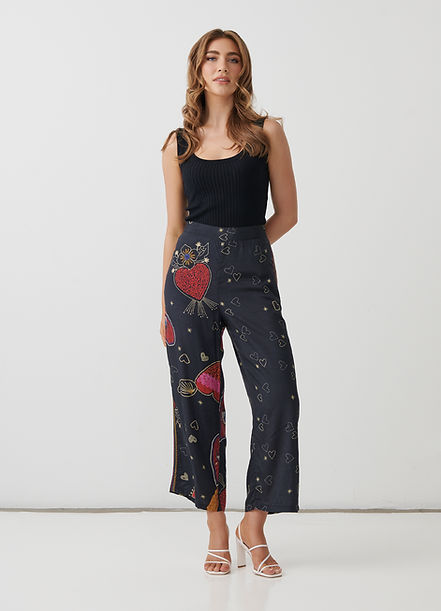 A woman modeling high-waisted cropped pants with a dark navy base and a colorful print of red hearts, planets, and small gold stars, paired with a black ribbed tank top and white strappy heels against a white studio background.