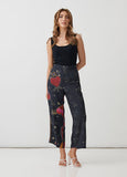 A woman modeling high-waisted cropped pants with a dark navy base and a colorful print of red hearts, planets, and small gold stars, paired with a black ribbed tank top and white strappy heels against a white studio background.