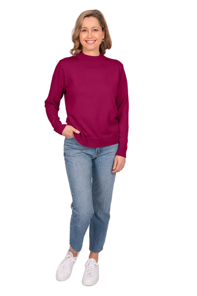 A woman modeling a long-sleeve mock neck sweater in deep magenta, paired with blue jeans and white sneakers, standing against a light gray studio background.