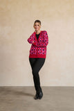 A woman modeling a bright pink cardigan with a bold magenta and purple circular pattern, paired with black pants and black boots, standing against a textured beige studio wall.