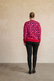 A woman viewed from behind wearing a vibrant red crewneck sweater with a circular black, pink, and purple pattern, paired with black pants and black ankle boots, standing against a textured beige wall.