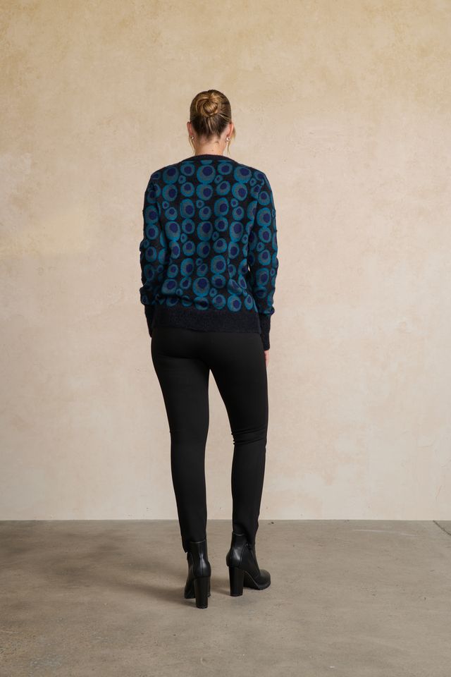 A woman viewed from behind wearing a dark blue and black patterned knit sweater, black leggings, and black ankle boots, standing against a textured beige wall.