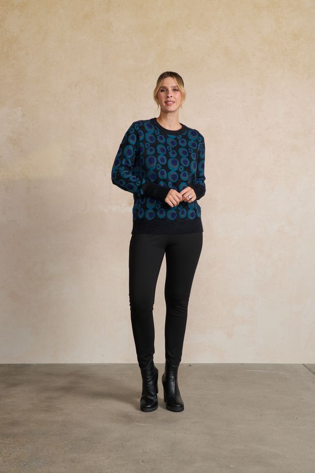 A woman modeling a dark teal and black long-sleeve knit sweater with a circular pattern, paired with black leggings and black ankle boots, standing against a textured beige plaster wall.