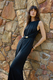 A woman wearing a sleeveless black pleated maxi dress with a twisted waist detail and a small metallic embellishment, posing against a rustic stone wall.