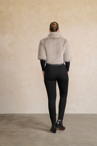 A model viewed from behind wearing a light beige shaggy faux-fur cropped jacket over a black long-sleeve top and black leggings, standing against a textured beige wall.