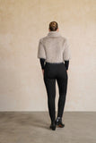 A model viewed from behind wearing a light beige shaggy faux-fur cropped jacket over a black long-sleeve top and black leggings, standing against a textured beige wall.