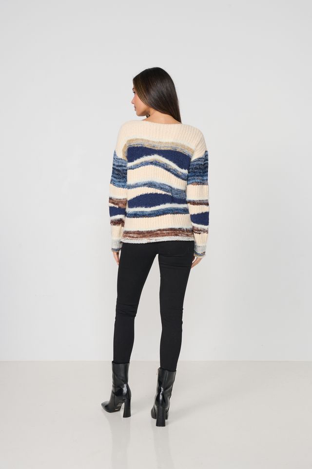 A woman viewed from the back wearing a cream knit sweater with horizontal blue, navy, and brown stripes, paired with black pants and black heeled boots, against a plain light gray studio background.