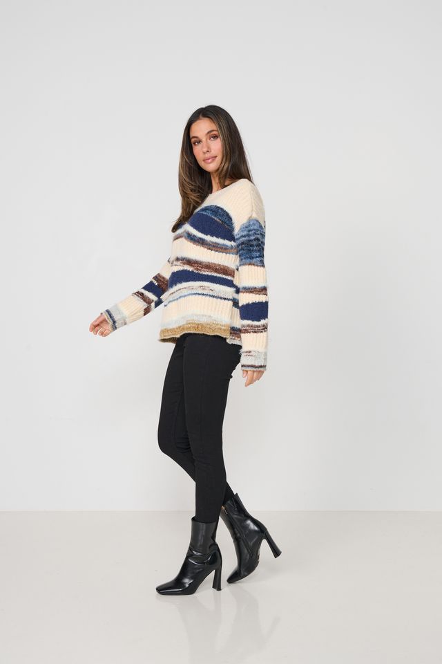 A woman modeling a fuzzy knit sweater with horizontal stripes in cream, blue, and brown, paired with black pants and black ankle boots, against a plain white studio background.