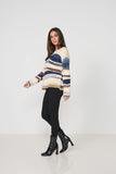 A woman modeling a fuzzy knit sweater with horizontal stripes in cream, blue, and brown, paired with black pants and black ankle boots, against a plain white studio background.