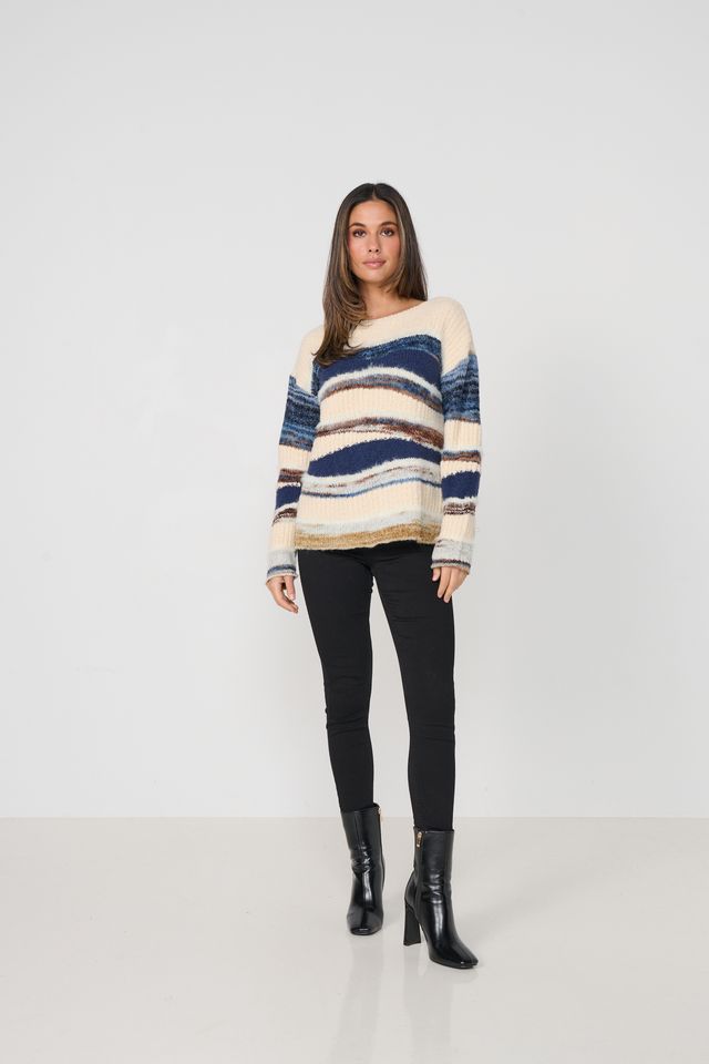 A woman modeling a fuzzy knit off-the-shoulder sweater with horizontal stripes in cream, navy blue, and brown, paired with black pants and black ankle boots, against a plain white studio background.