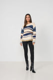 A woman modeling a fuzzy knit off-the-shoulder sweater with horizontal stripes in cream, navy blue, and brown, paired with black pants and black ankle boots, against a plain white studio background.