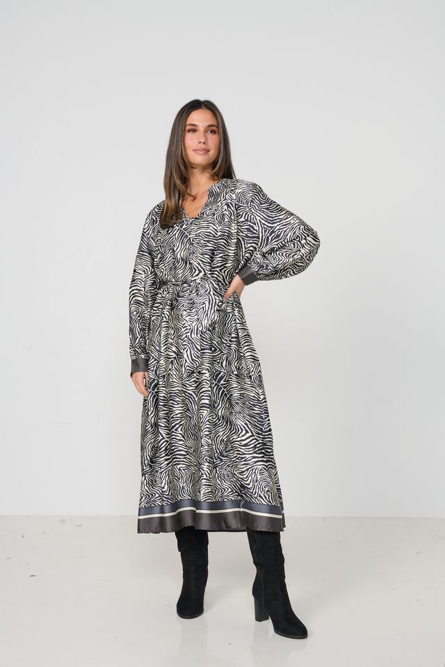 A woman modeling a long-sleeved midi dress with a black and white zebra-like abstract print, paired with black knee-high boots, against a plain white studio background.