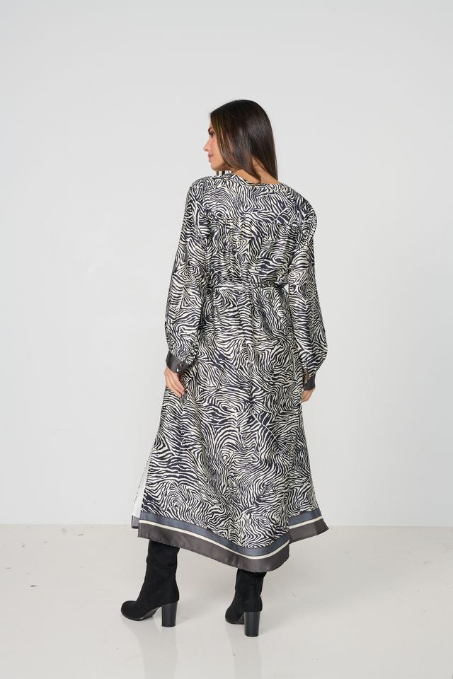A woman viewed from the back wearing a long-sleeved, knee-length tunic dress with a black and white zebra-like abstract print and a dark gray border, paired with black ankle boots, against a plain white studio background.
