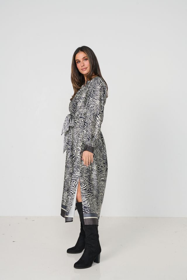 A woman modeling a long-sleeved wrap-style midi dress with a black and white zebra print, paired with black knee-high boots, against a plain white studio background.
