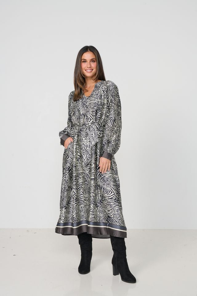 A woman modeling a long-sleeved midi dress with a black and white zebra-like abstract pattern, paired with black boots, against a plain white studio background.