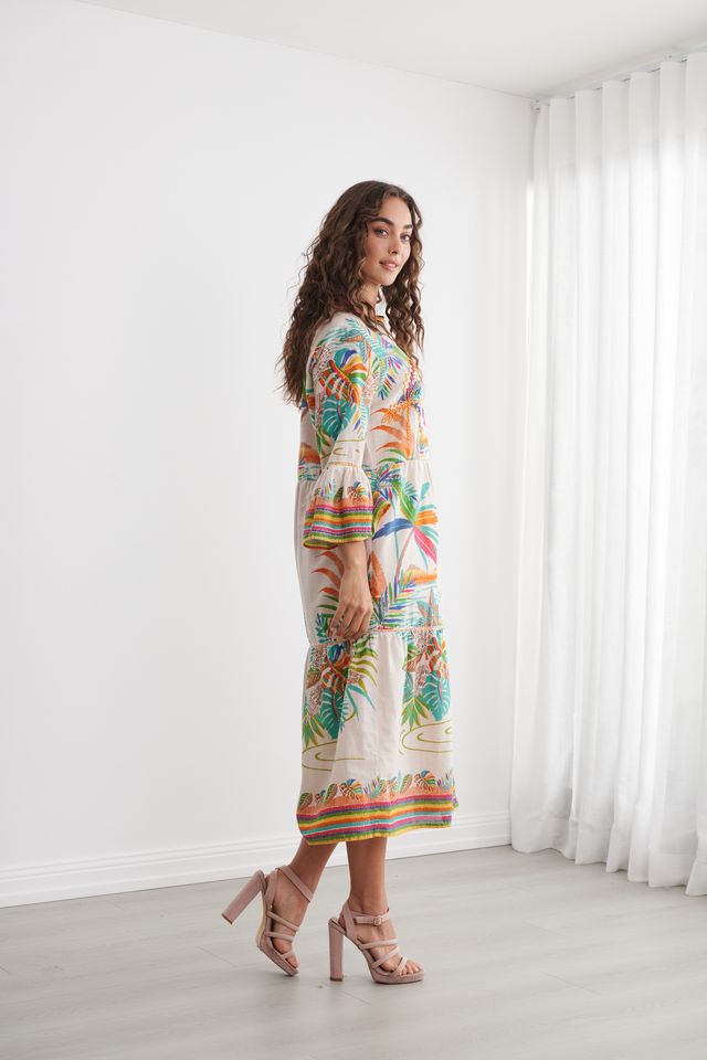 A woman modeling a long-sleeve midi dress with a colorful tropical beach print, standing in a bright, minimalist room with white walls and sheer curtains.