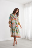 A woman modeling a tropical print midi dress with three-quarter sleeves in a bright, modern room with white walls and sheer curtains.