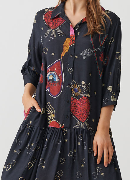 A woman wearing a dark navy blue button-up shirt dress with a whimsical red, gold, and white pattern of hearts, stars, and abstract shapes, shown against a light gray background.