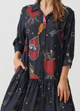 A woman wearing a dark navy blue button-up shirt dress with a whimsical red, gold, and white pattern of hearts, stars, and abstract shapes, shown against a light gray background.
