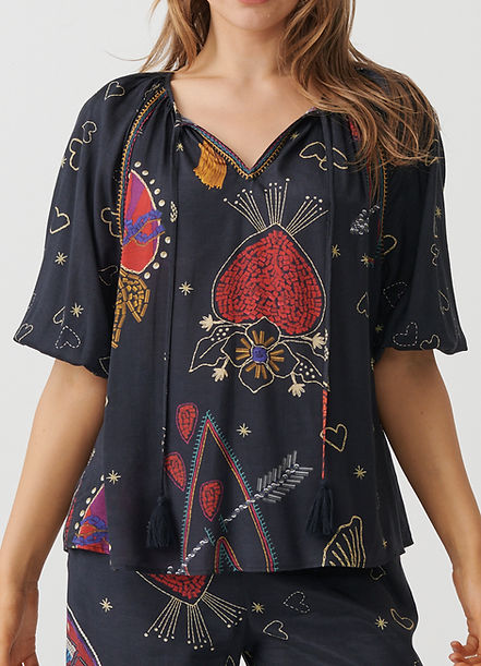 A woman wearing a black short-sleeve blouse with a colorful embroidered pattern of strawberries, flowers, hearts, and stars, paired with matching black pants.