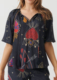 A woman wearing a black short-sleeve blouse with a colorful embroidered pattern of strawberries, flowers, hearts, and stars, paired with matching black pants.