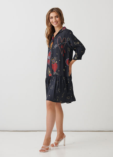 A woman modeling a short navy blue dress with a red and gold heart pattern, paired with white high-heeled sandals, set against a plain white studio background.
