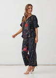 A woman modeling a matching two-piece lounge set featuring a short-sleeve top and wide-leg pants in navy blue with a colorful butterfly and gold heart pattern, paired with white strappy high-heeled sandals against a plain white studio background.