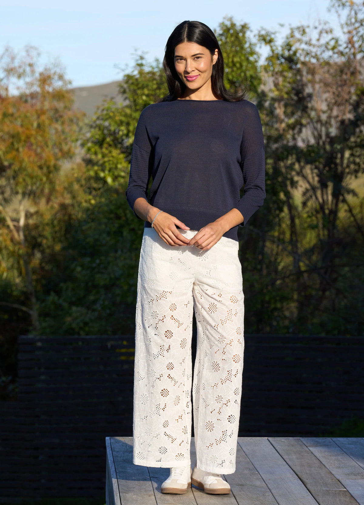 Woman wearing a navy top and white lace pants standing outdoors with trees in the background