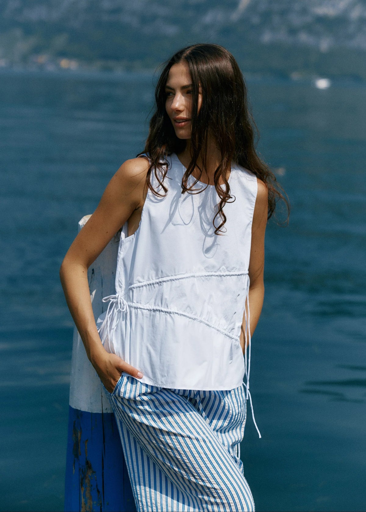 Woman in a white sleeveless top and blue striped pants standing by a body of water.