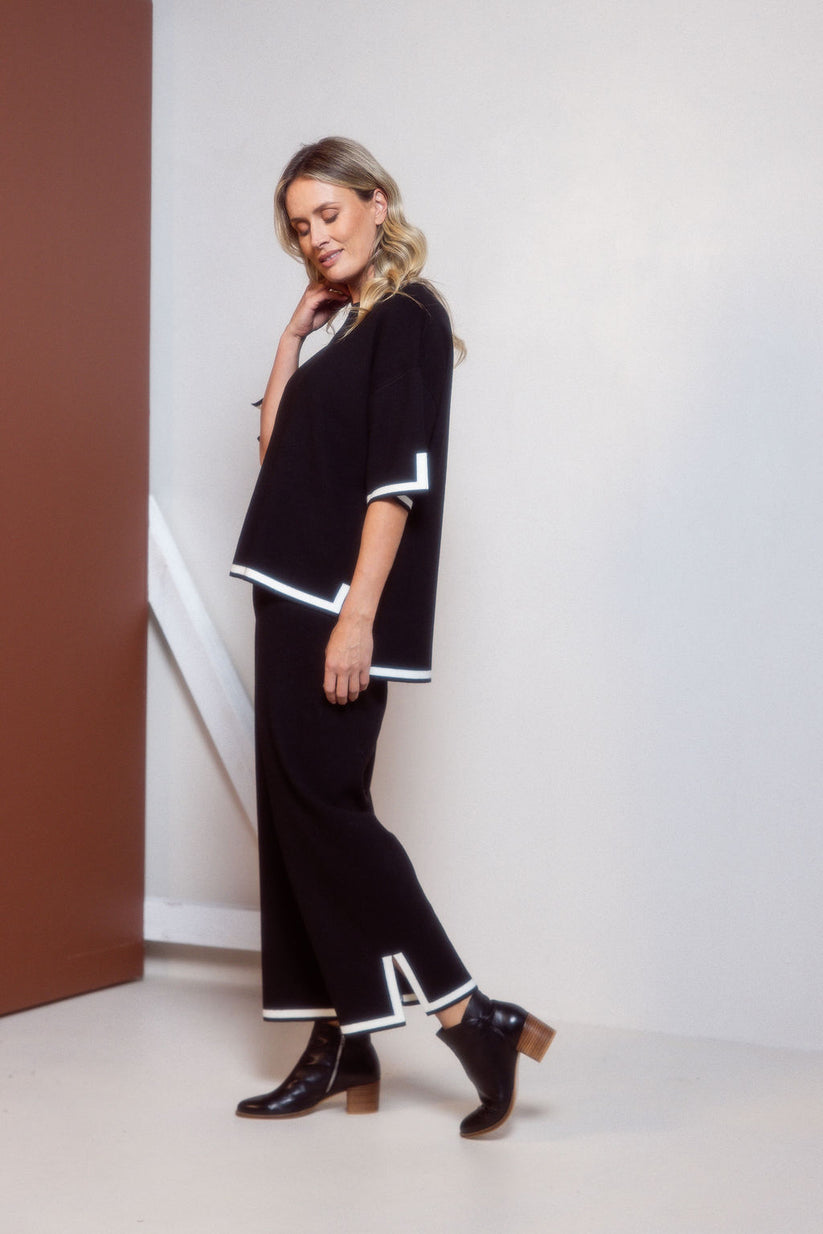 Woman wearing a black outfit with white trim standing against a white wall.